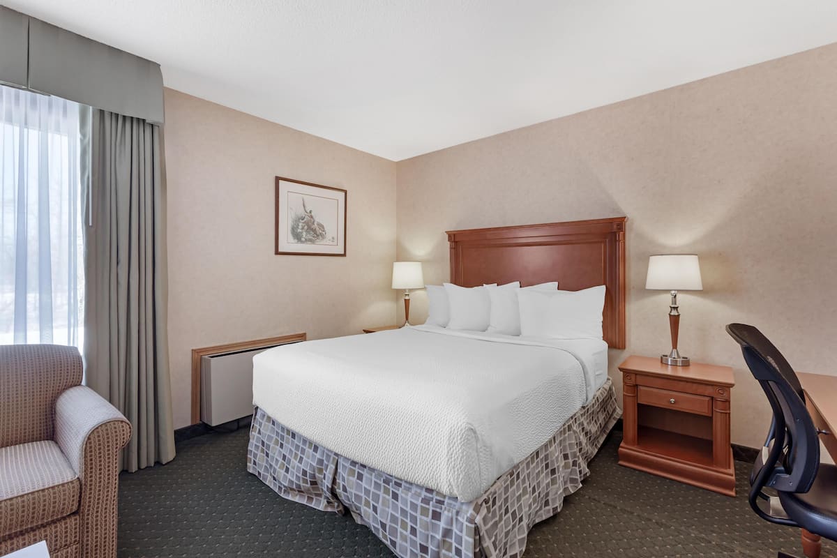 Ein Hotelzimmer mit einem großen Bett, zwei Nachttischen, einem Sessel, einem Schreibtisch und einem fenster mit Vorhängen.