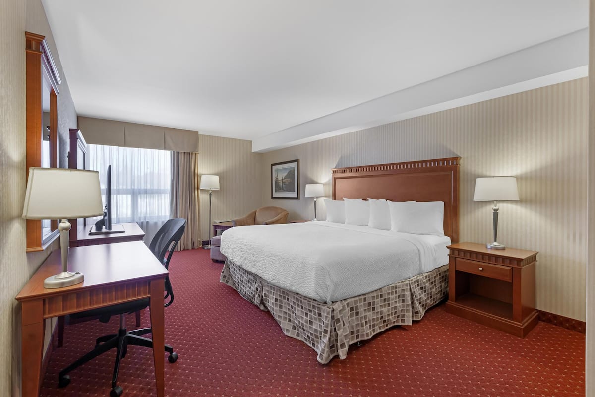 Ein Hotelzimmer mit einem großen Bett, einem Schreibtisch mit Stuhl, einem kleinen Tisch mit Lampe und einem Fenster mit Vorhängen.