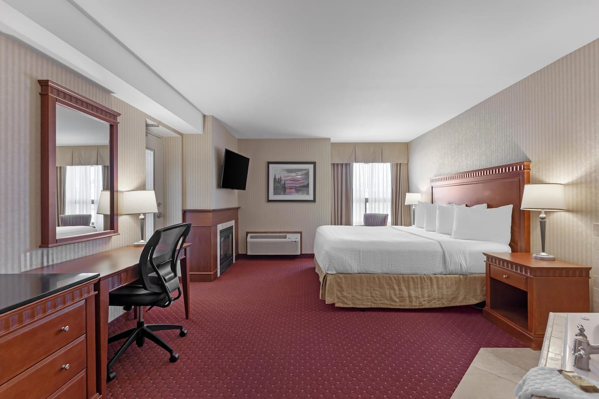 Ein Hotelzimmer mit einem großen Bett, einem Schreibtisch mit Stuhl, einem Schminktisch, einem Fernseher und einem Fenster mit Vorhängen.