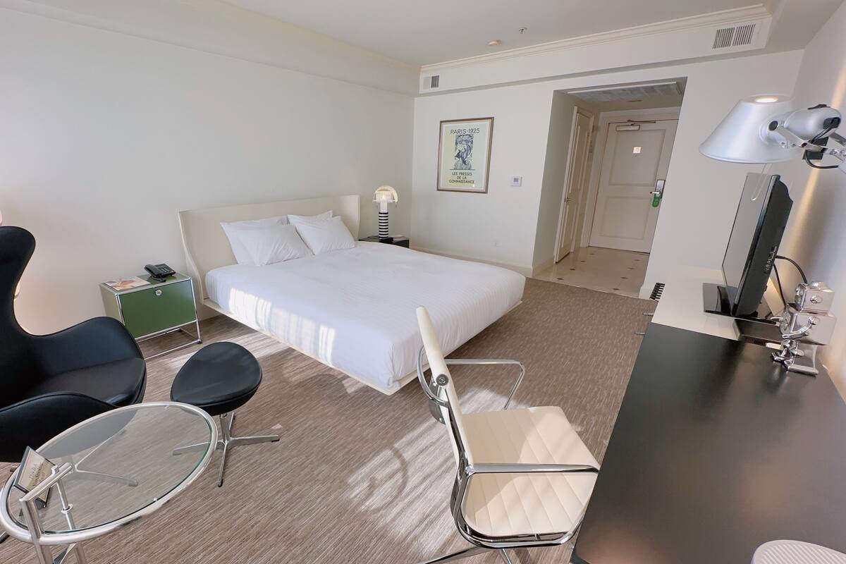Habitación de hotel con cama, escritorio, silla, mesita, lámpara y un cuadro en la pared.