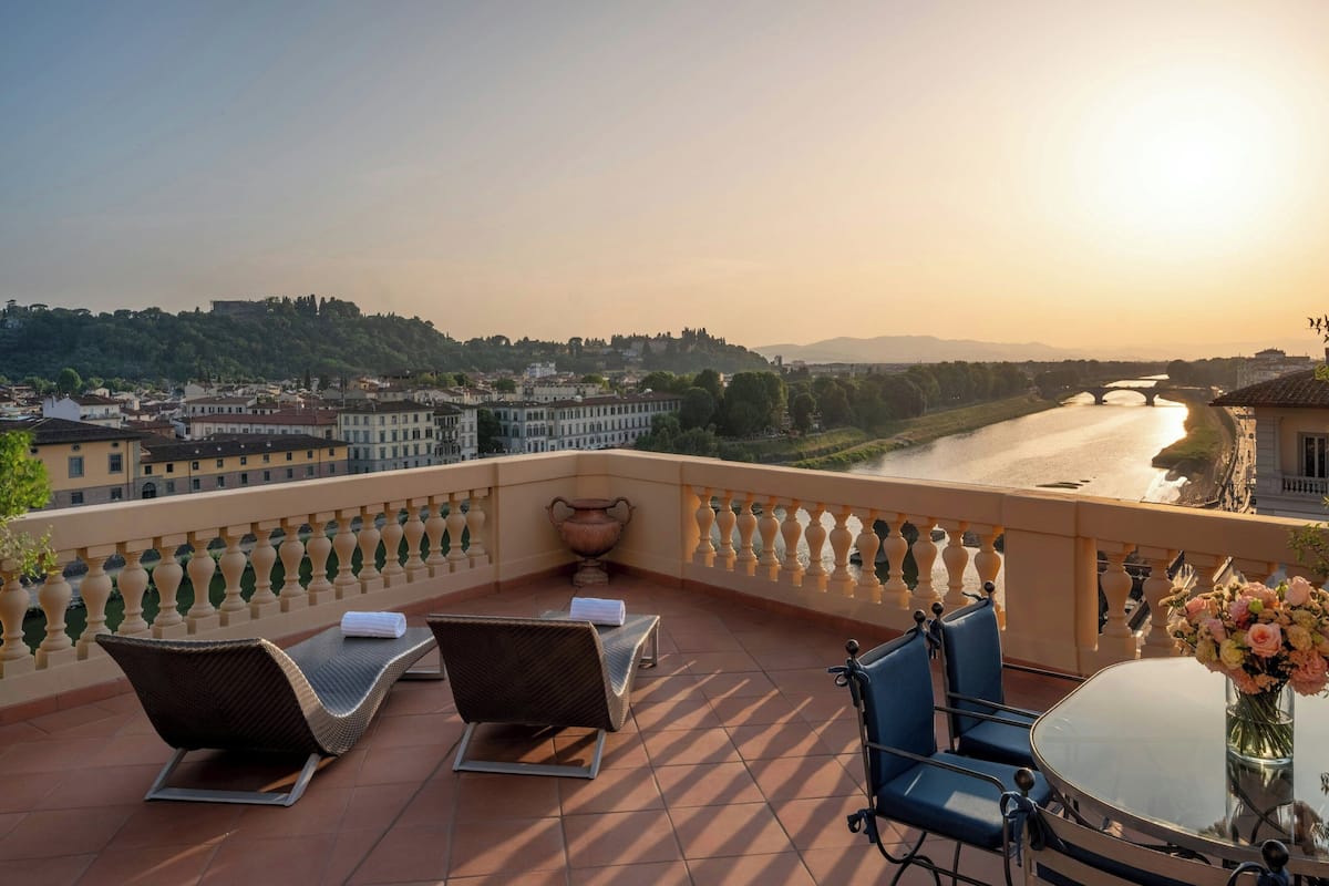 A terrace with lounge chairs, a table with a vase of flowers, and a view of a river and bridge.