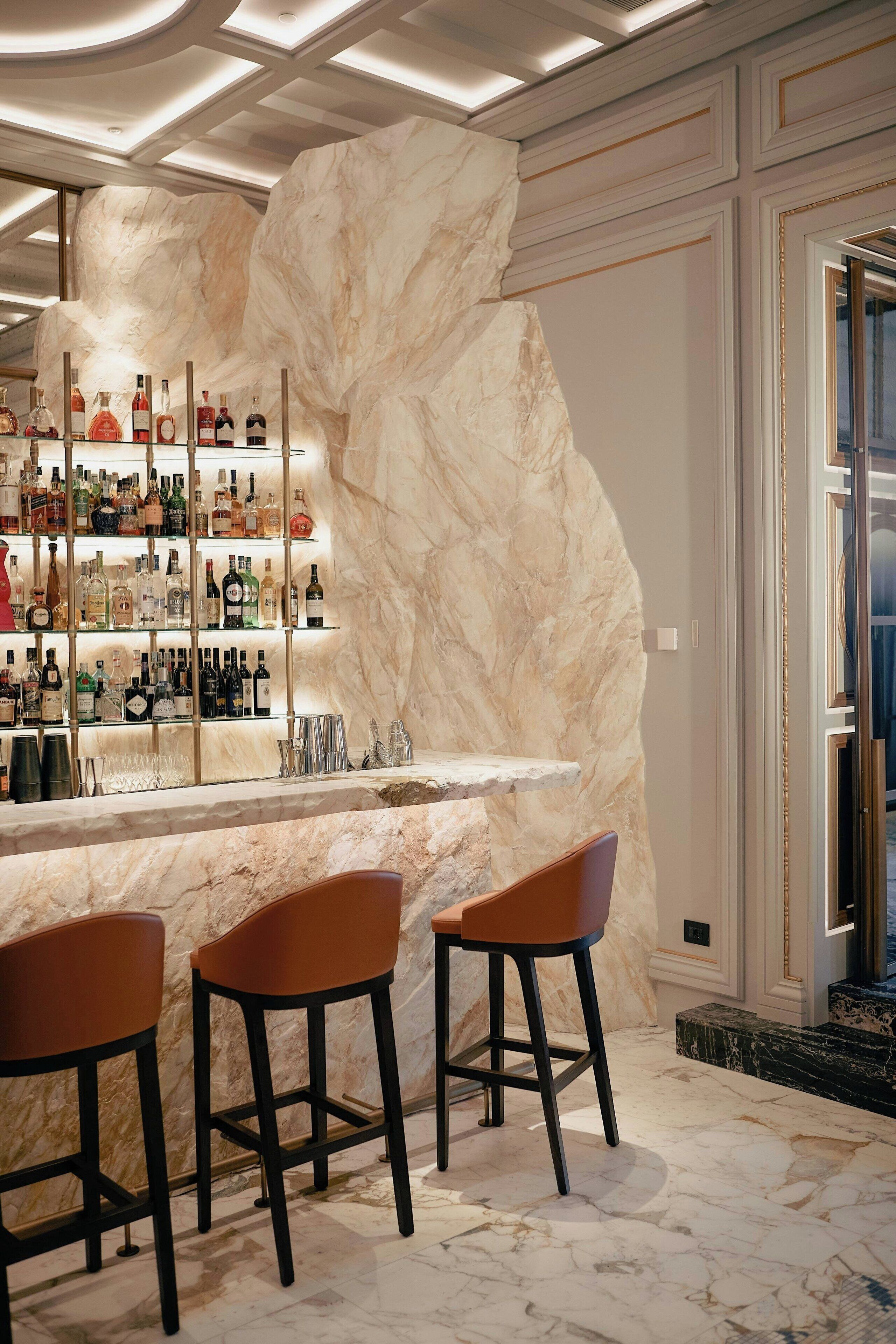 Una zona de bar con paredes de mármol, una variedad de botellas de licor y tres taburetes de bar.