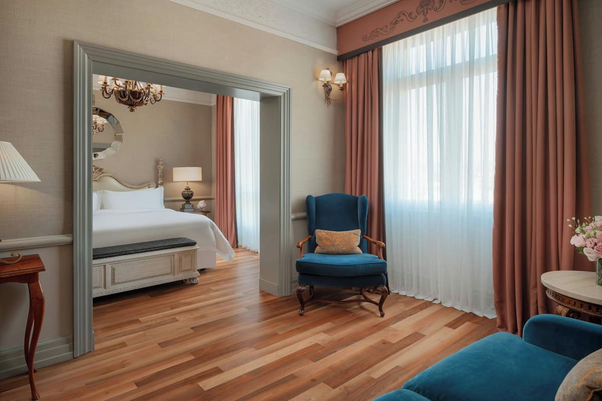 A hotel room with a bed, a blue armchair, a round table with a vase, and a large window with curtains.