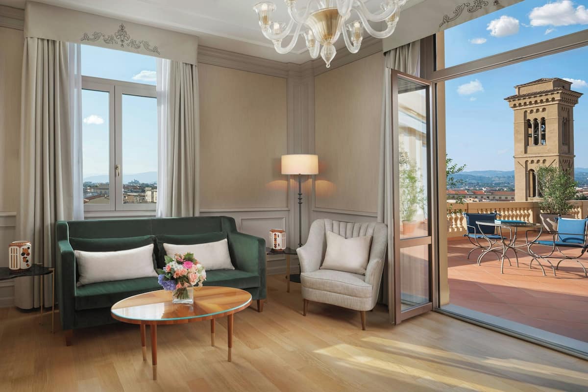 A spacious room with a green sofa, a beige armchair, a round coffee table, and a lamp. A large window offers a view of a tower and a cityscape.
