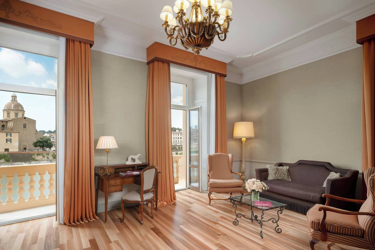 A spacious room with a wooden floor, a desk with a lamp, a sofa, and a chair. There is a view of a building outside through large windows.