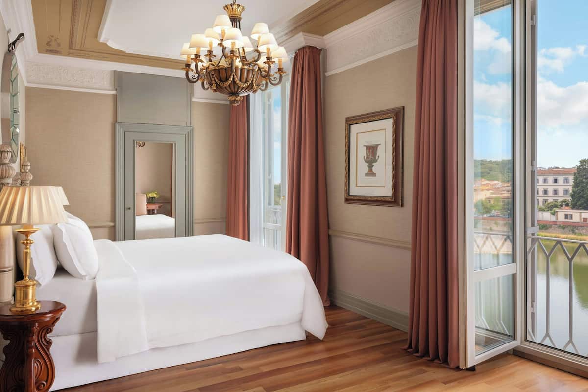 A hotel room with a large bed, a chandelier, a view of a cityscape through a balcony, and a framed picture on the wall.