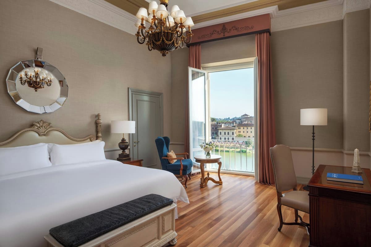 A hotel room with a large bed, a chandelier, a round mirror, a desk with a lamp, a chair, and a view of a cityscape through a large window.