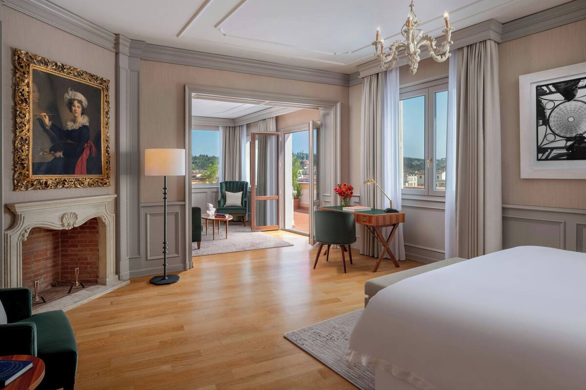 A spacious hotel room with a large bed, a fireplace, a desk with a chair, and a balcony with a view of the city.