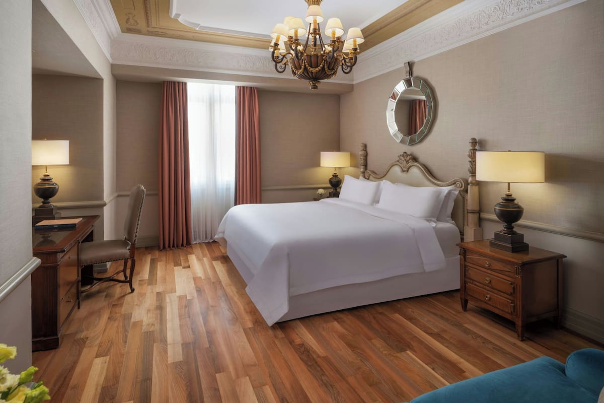 A hotel room with a large bed, wooden flooring, a chandelier, a desk with a lamp, and a chair.