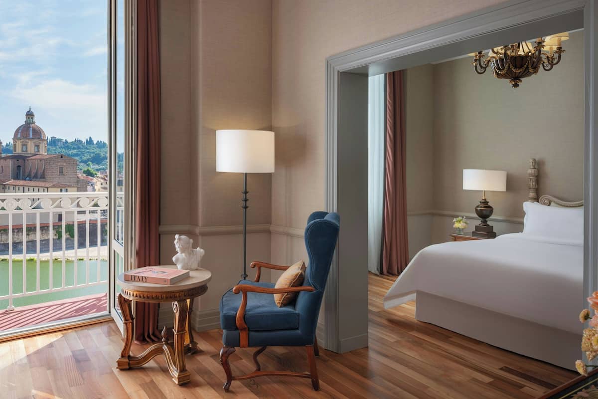 A hotel room with a large bed, a sitting area with a chair and table, and a view of a cityscape through large windows.