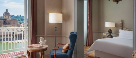 A hotel room with a large bed, a sitting area with a chair and table, and a view of a cityscape through large windows.