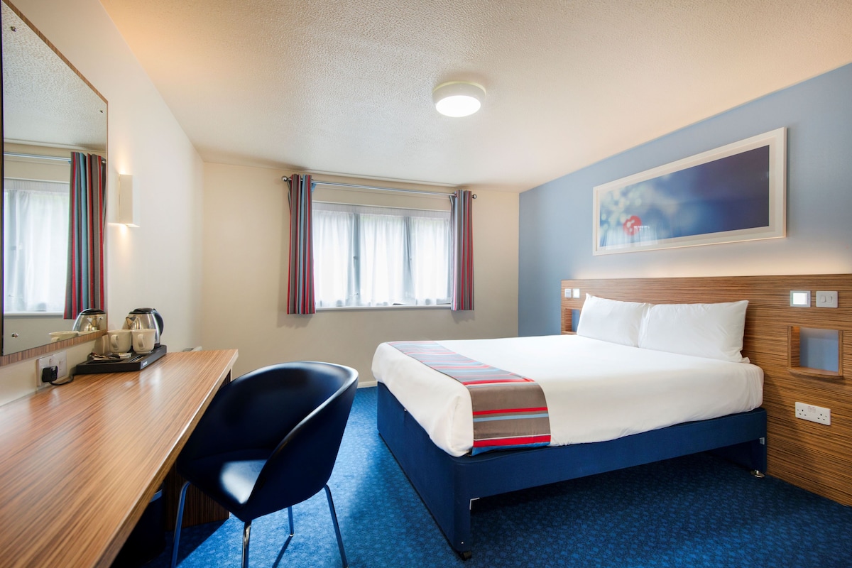 Habitación de hotel con cama, escritorio, silla, ventana con cortinas y un cuadro en la pared.