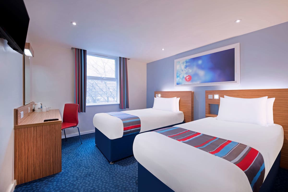 A hotel room with two beds, a desk, a chair, and a window with curtains.