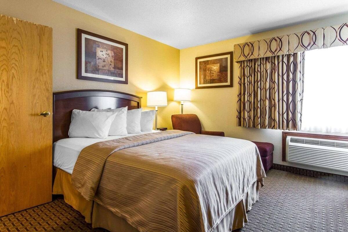 A hotel room with a bed, two bedside lamps, a chair, a window with curtains, and a wall-mounted air conditioner.