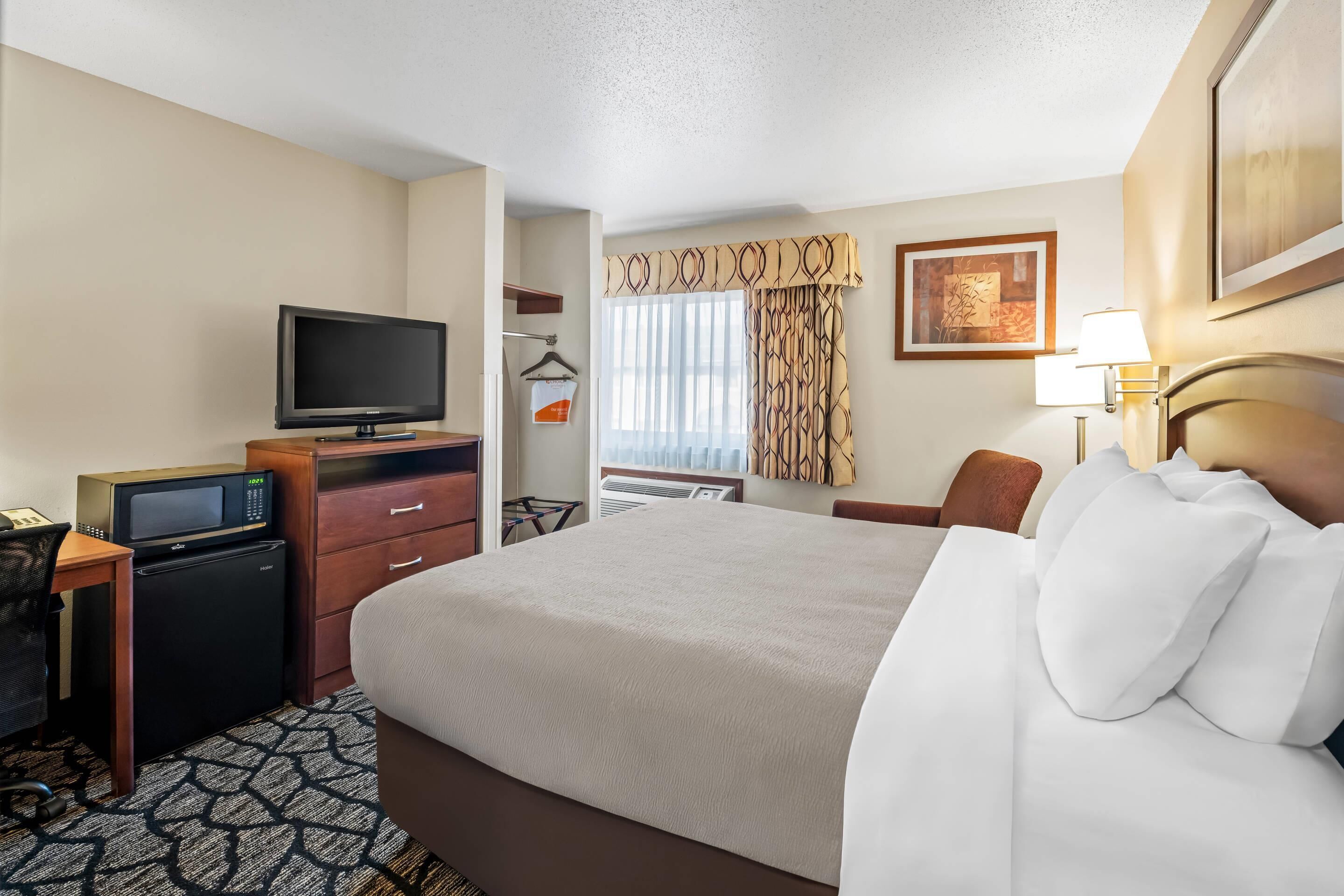 A hotel room with a bed, a desk with a television, a refrigerator, a chair, and a window with curtains.