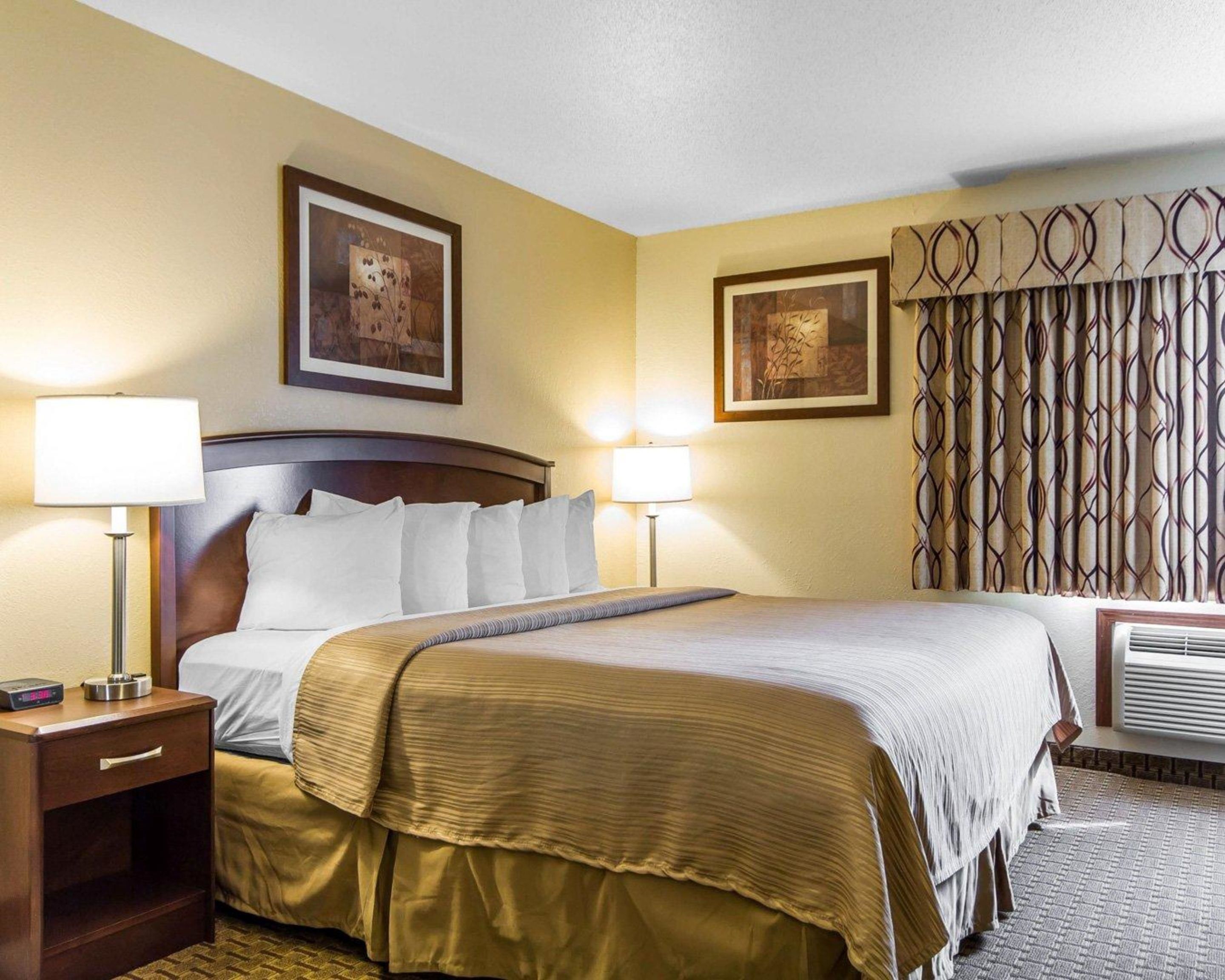 A hotel room with a large bed, two bedside lamps, a nightstand, a framed picture on the wall, a patterned curtain, and a window air conditioning unit.