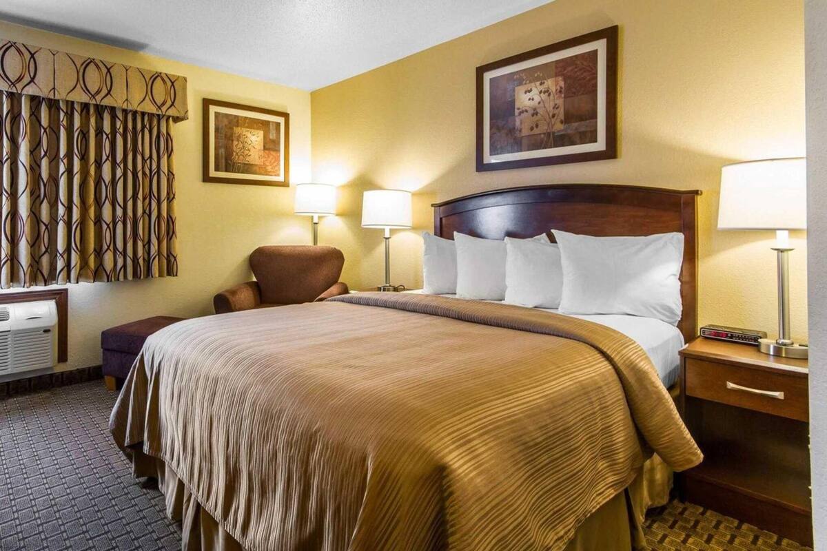 A hotel room with a large bed, two bedside lamps, a chair, a desk, and a wall-mounted air conditioner.