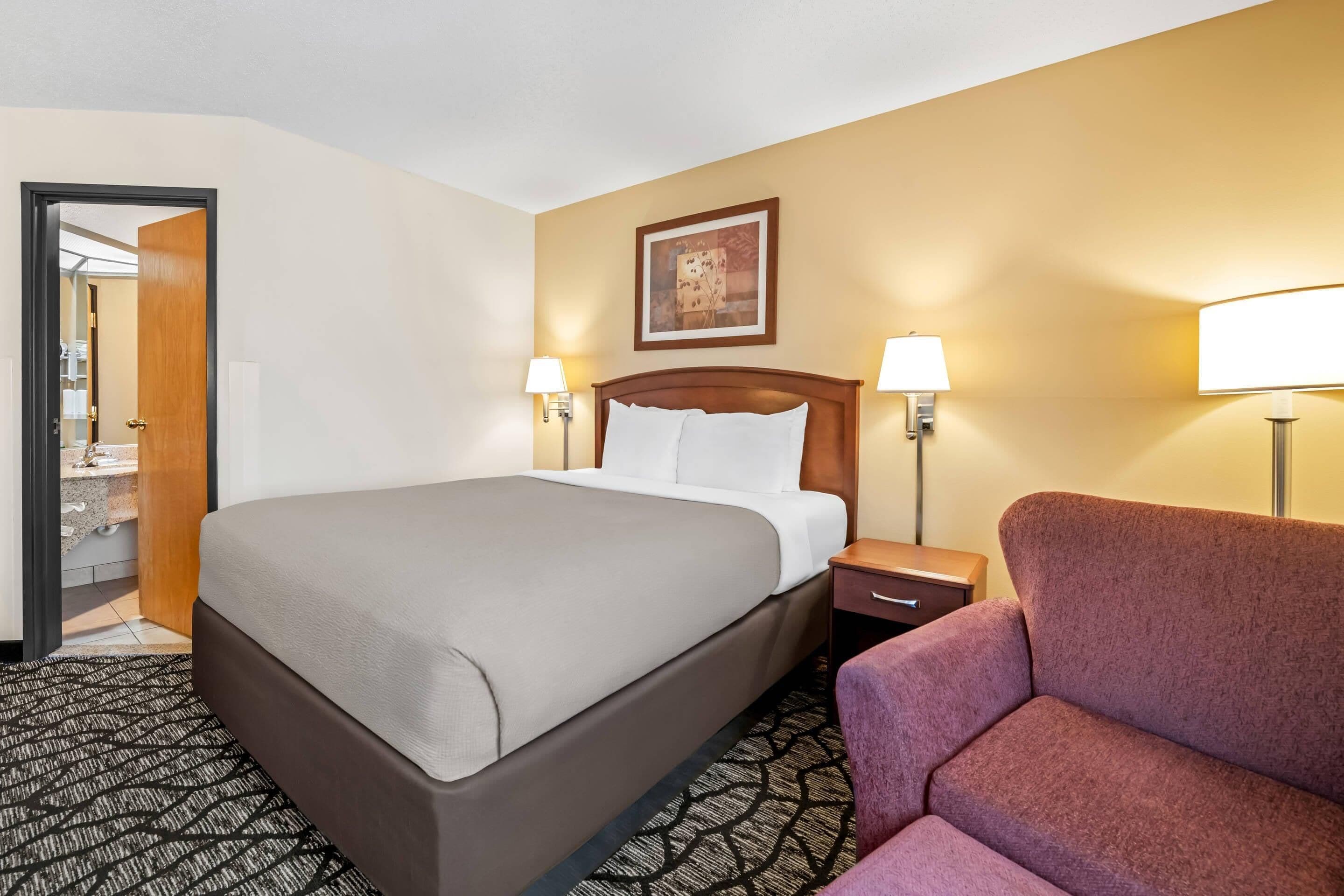 A hotel room with a large bed, two bedside lamps, a nightstand, a chair, and a door leading to a bathroom.