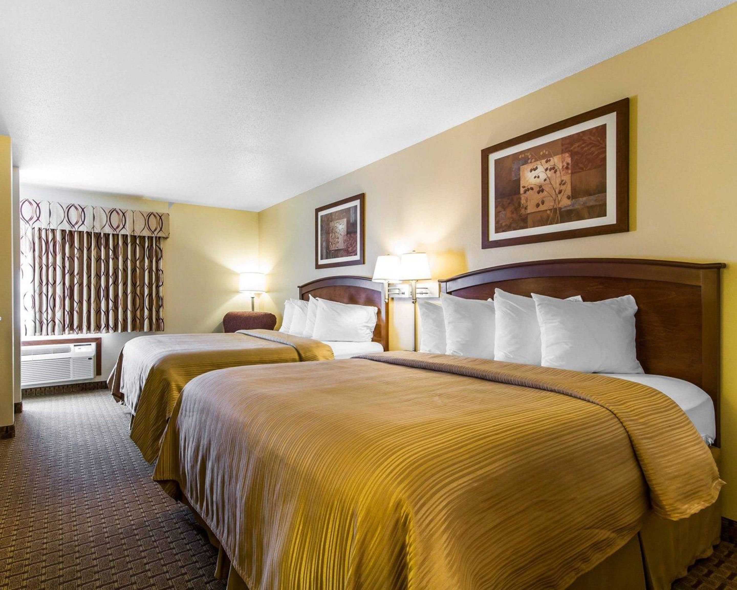 A hotel room with two beds, a large window with curtains, a wall-mounted artwork, and a lamp.