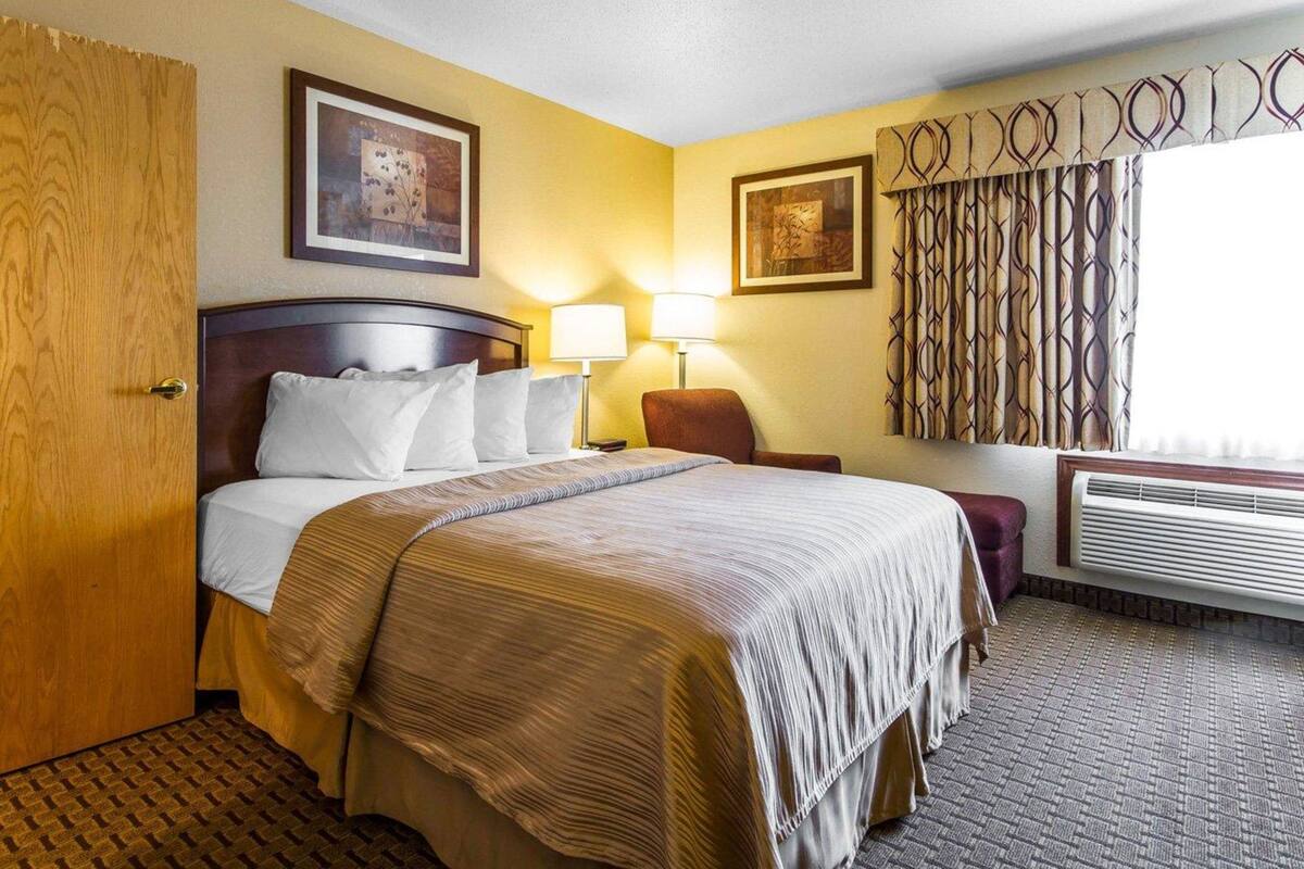 A hotel room with a bed, two bedside lamps, a chair, a window with curtains, and an air conditioning unit.