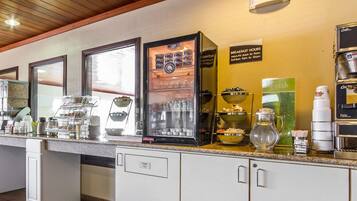 Un espace dédié au petit-déjeuner, avec un système de libre-service comprenant une machine à café, un présentoir de fruits et un panneau indiquant les heures du petit-déjeuner.
