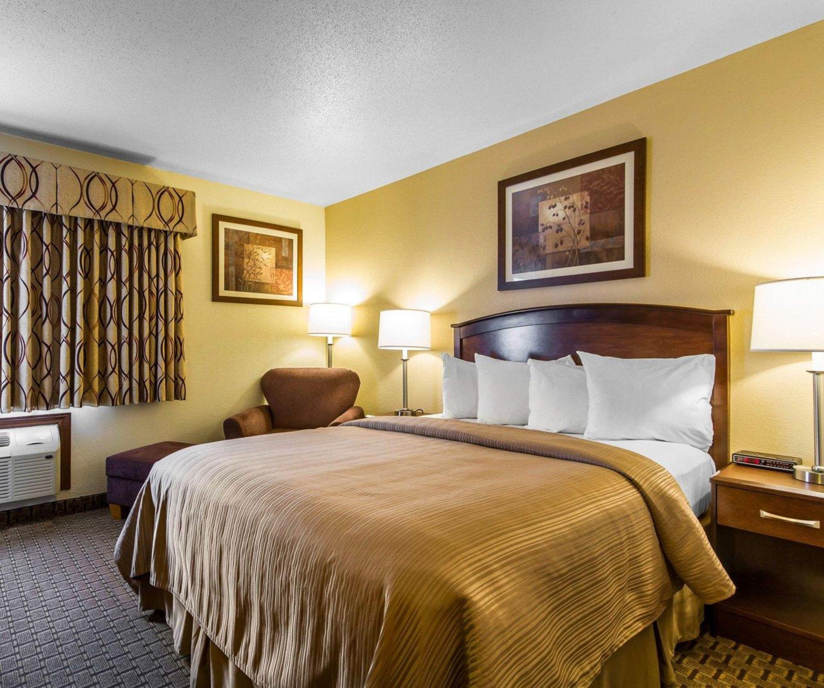 A hotel room with a large bed, two bedside lamps, a chair, a desk with a computer, and a wall-mounted air conditioner.