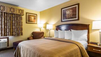 A hotel room with a large bed, two bedside lamps, a chair, a desk with a computer, and a wall-mounted air conditioner.