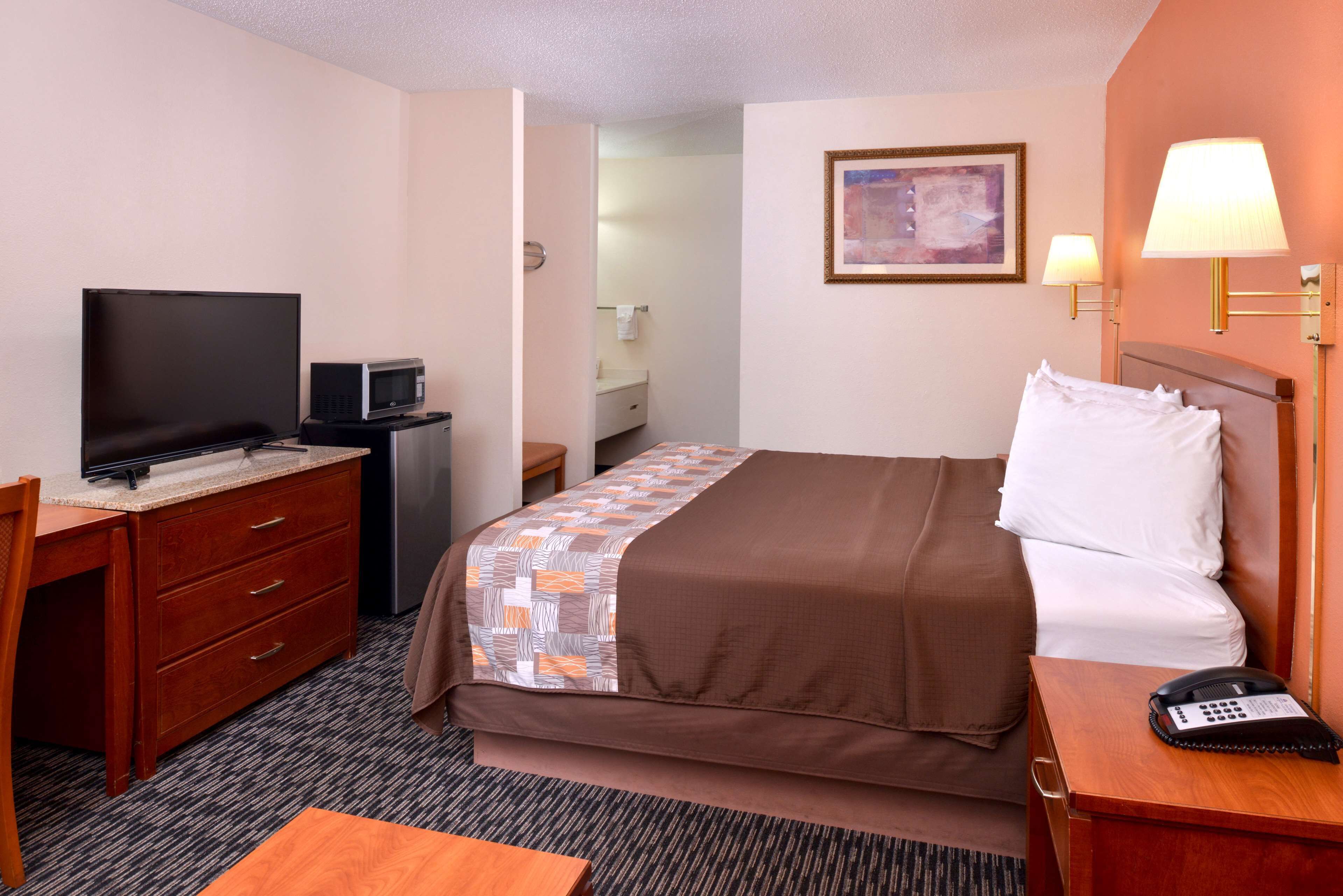 Une chambre d’hôtel comprenant un lit, un bureau avec une télévision et un micro-ondes, une table de chevet avec une lampe, et une salle de bain visible à travers une porte ouverte.
