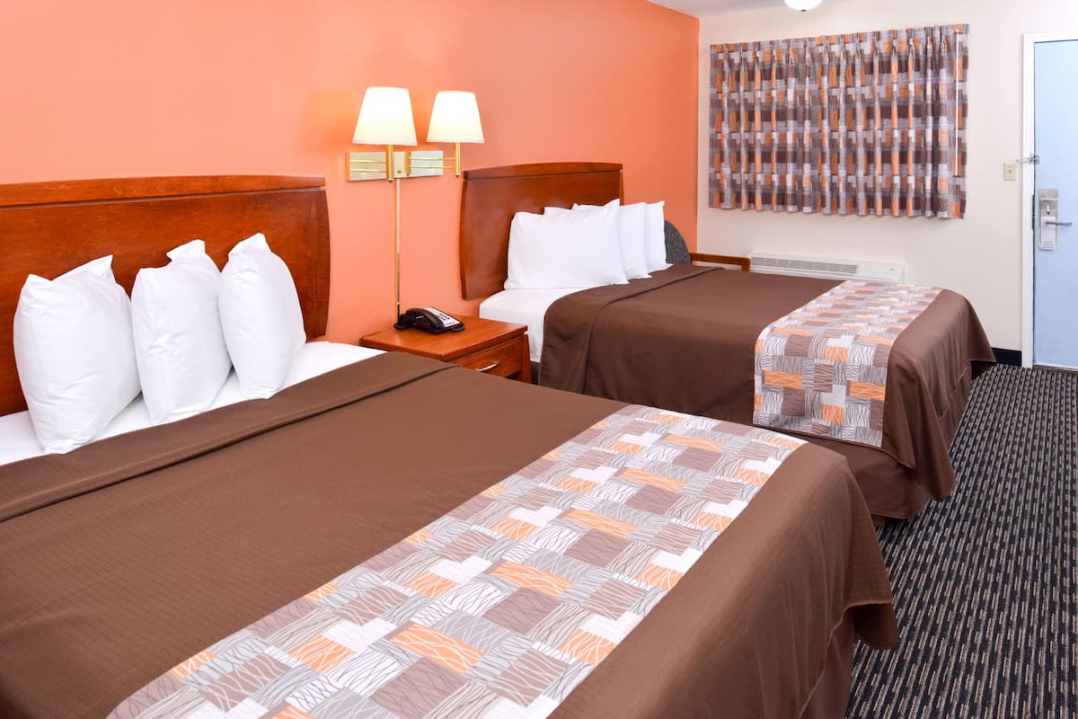 A hotel room with two beds, each with white and brown bedding, a nightstand with a phone, and a wall-mounted lamp.