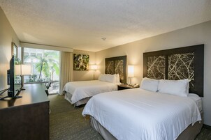 Habitación de hotel con dos camas, un escritorio, televisor y balcón con vista a palmeras.
