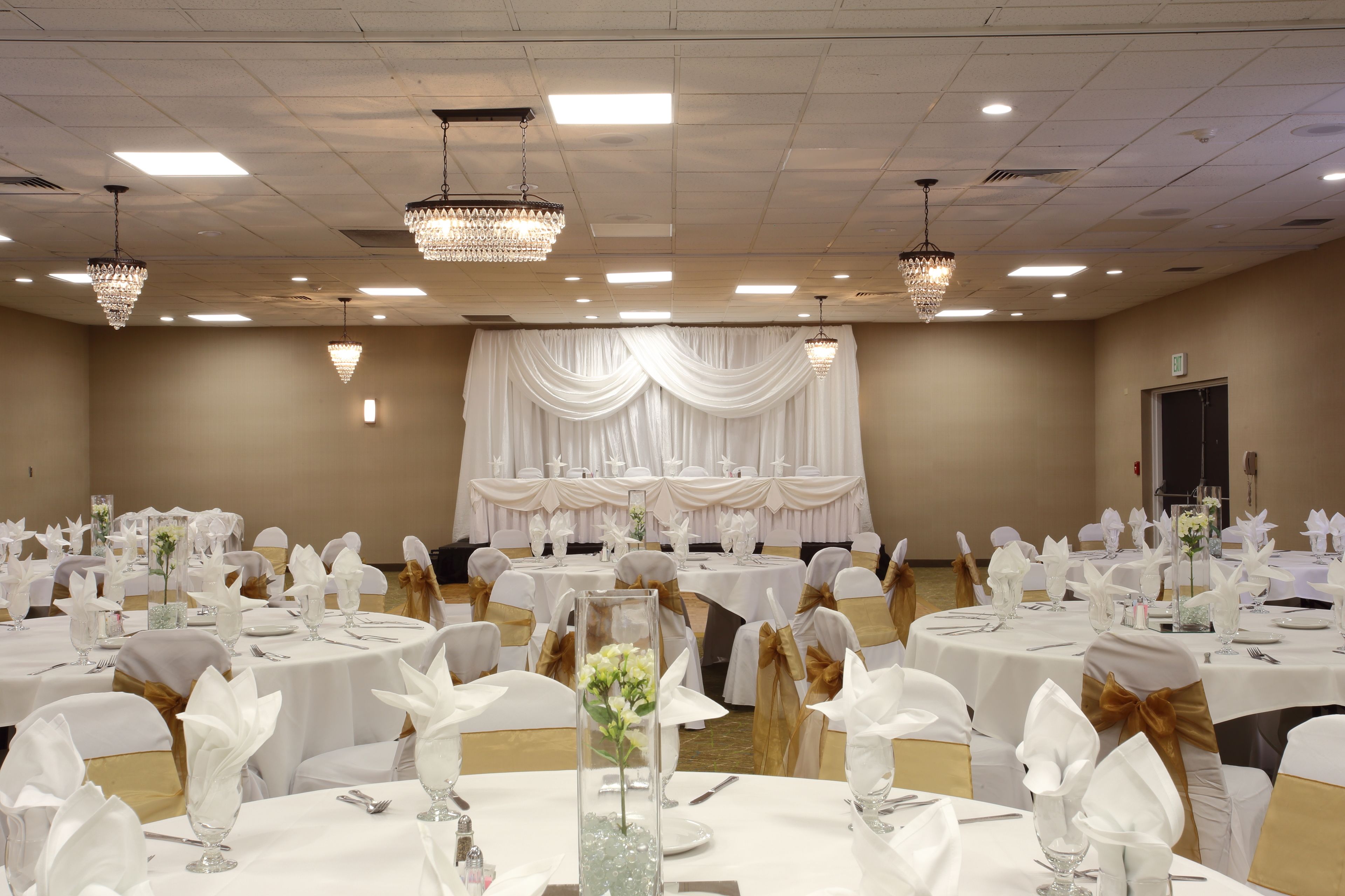 Uma sala de banquetes com mesas redondas preparadas para um evento formal, com toalhas brancas, fitas douradas e lustres de cristal.