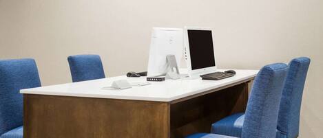 A conference room with a white table, two blue chairs, a white desk, a desktop computer, a monitor, a keyboard, and a mouse.