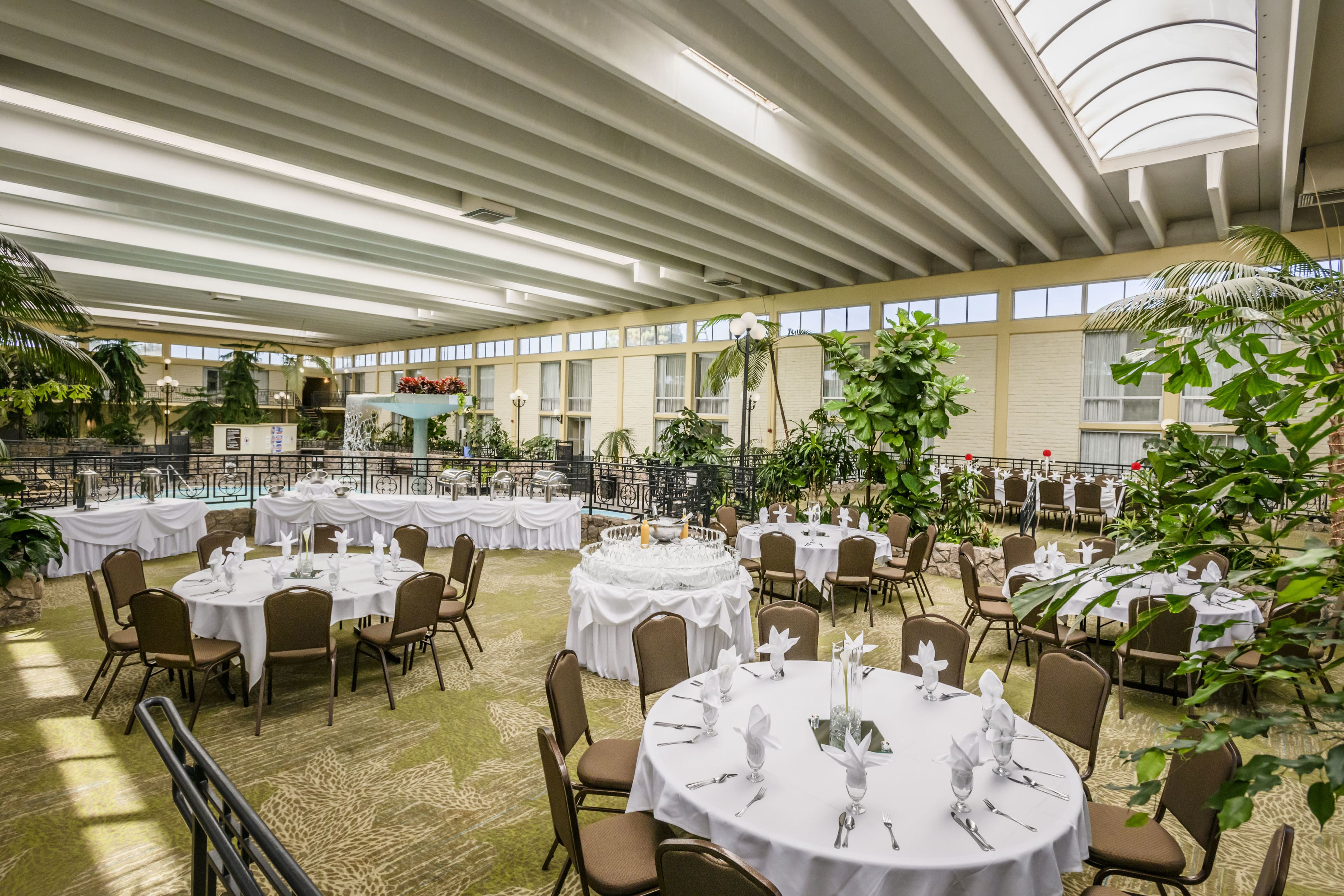 A spacious indoor dining area with round tables set for a formal event, surrounded by chairs and potted plants.