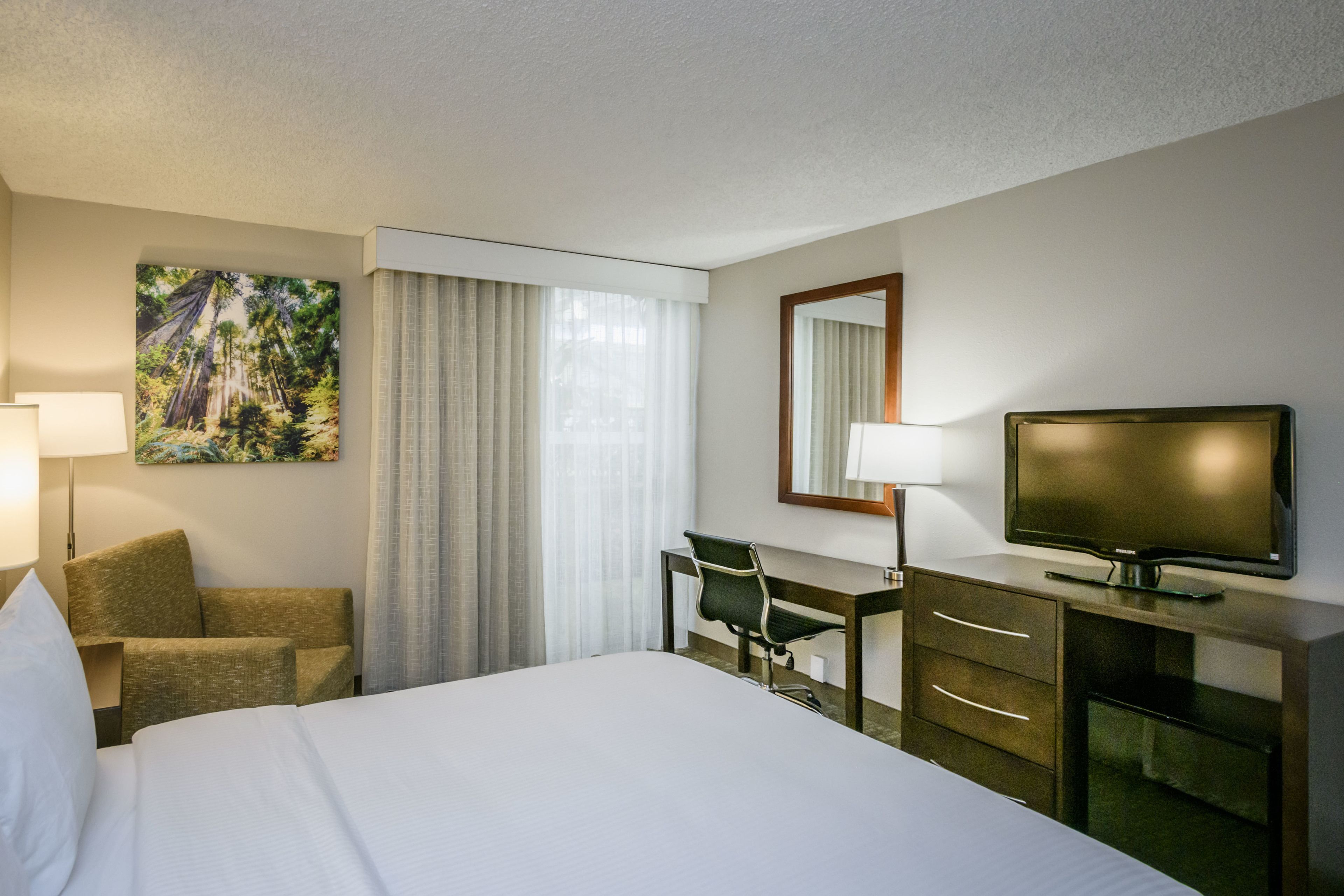Habitación de hotel con cama, escritorio, silla, televisión, espejo y ventana con cortinas.