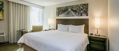 A hotel room with a large bed, two bedside tables, a chair, a window with curtains, a wall-mounted painting, and a wall-mounted heater.
