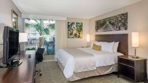 A hotel room with a bed, a desk with a TV, a chair, a nightstand, and a balcony with a view of palm trees. - Wyndham Garden Fresno Yosemite Airport (Fresno)