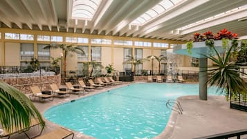 An indoor swimming pool with lounge chairs and palm trees, surrounded by large windows and a high ceiling.
