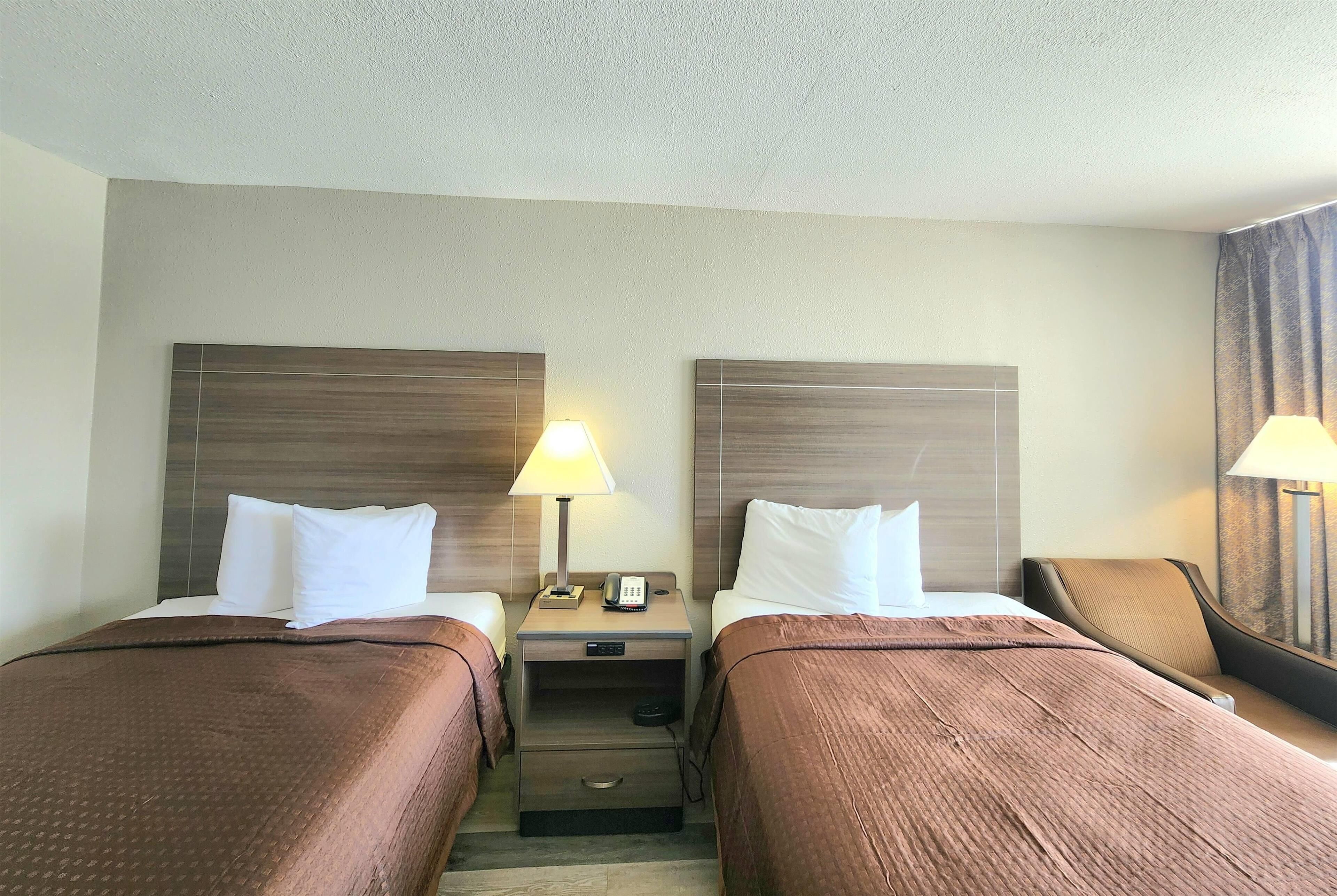 Une chambre d’hôtel avec deux lits, chacun recouvert d’une couverture marron et agrémenté d’oreillers blancs. Une table de chevet se trouve entre les deux lits, avec une lampe et un téléphone.