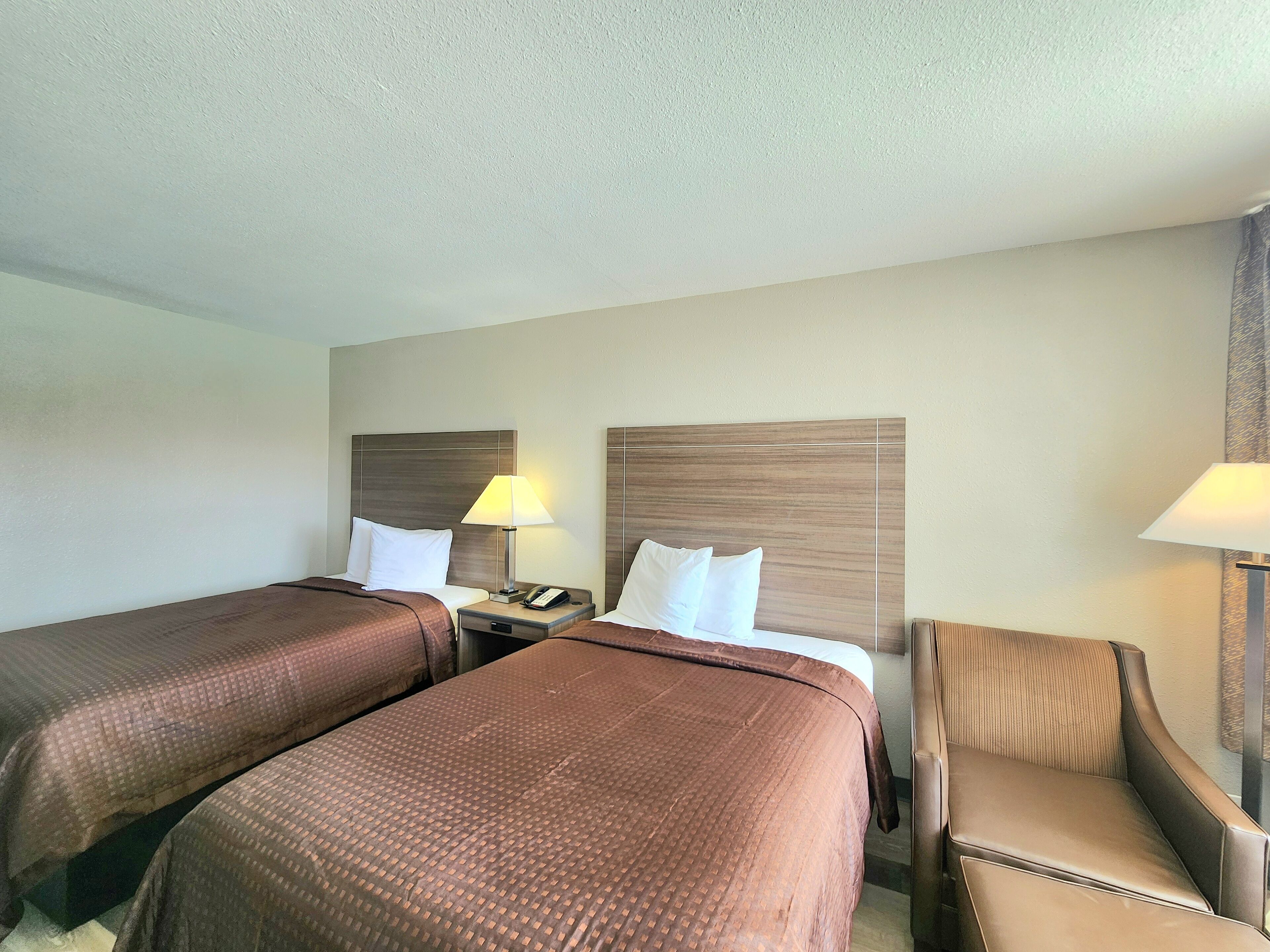 A hotel room with two beds, a nightstand, a lamp, and a chair.