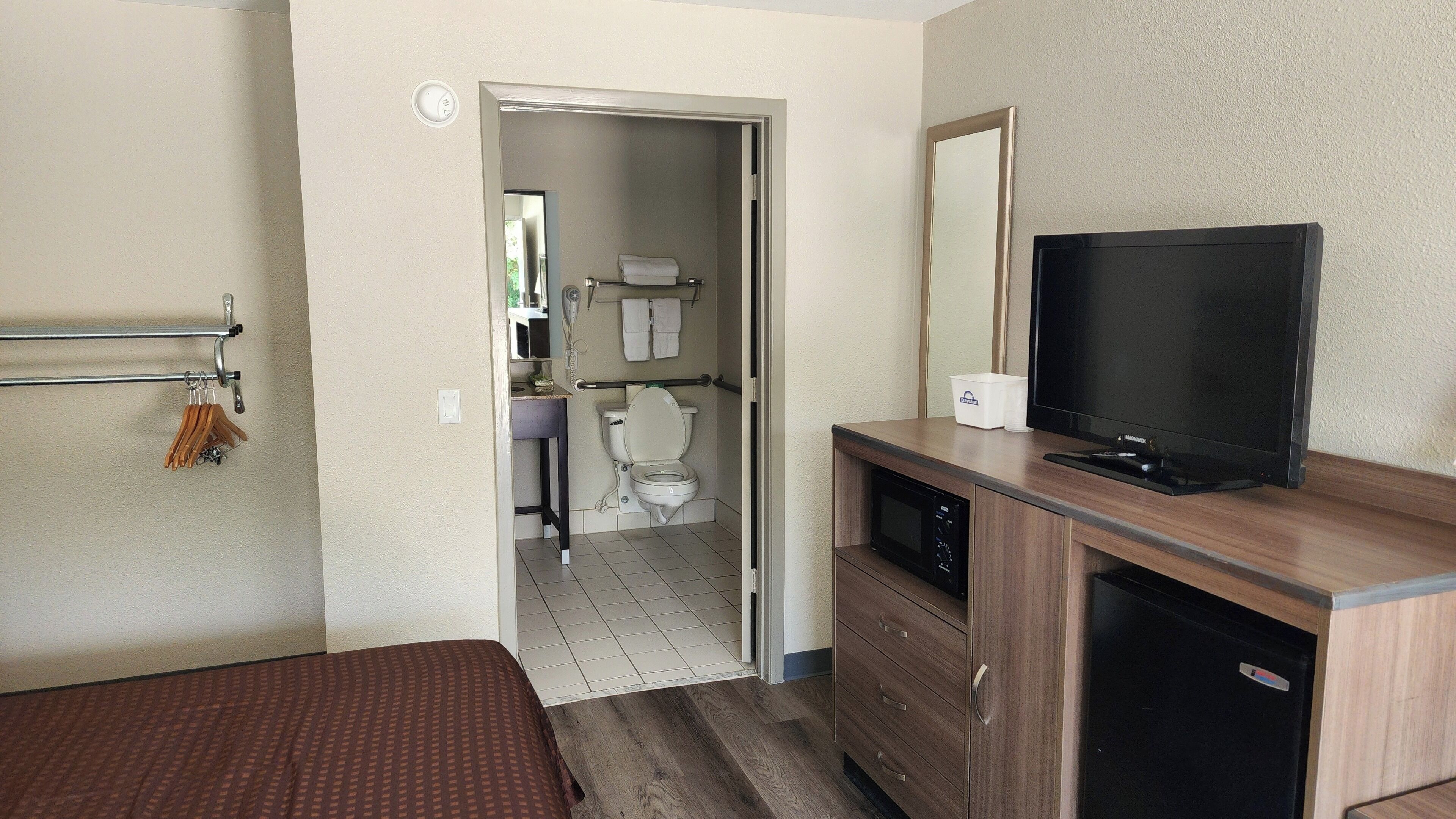 A hotel room with a bed, a TV, a wooden dresser, a microwave, and a bathroom visible through an open door.