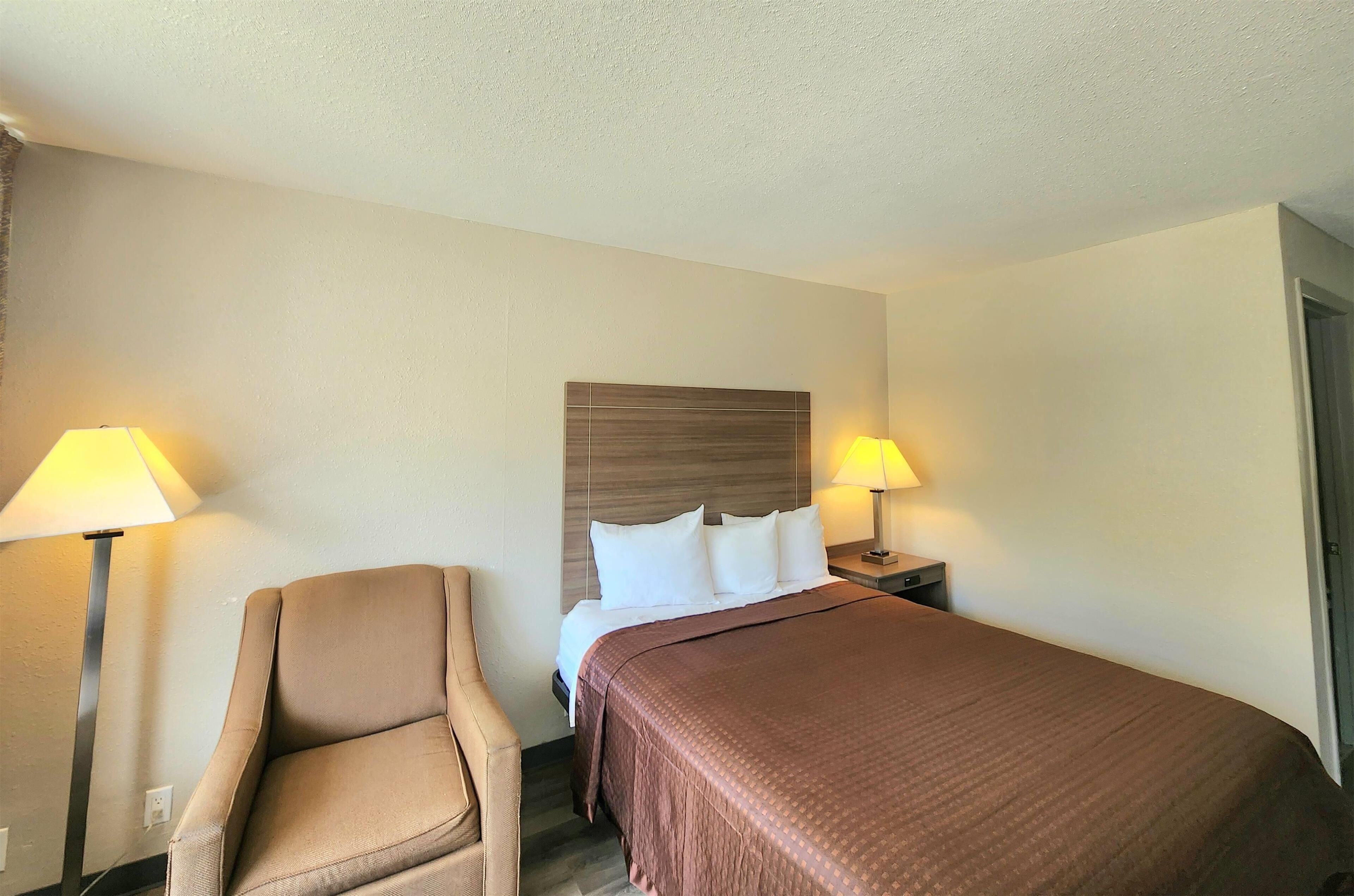 Quarto de hotel com cama, duas luminárias de cabeceira, uma cadeira e painel de cabeceira em madeira.