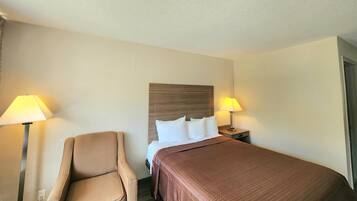 Quarto de hotel com cama, duas luminárias de cabeceira, uma cadeira e painel de cabeceira em madeira.