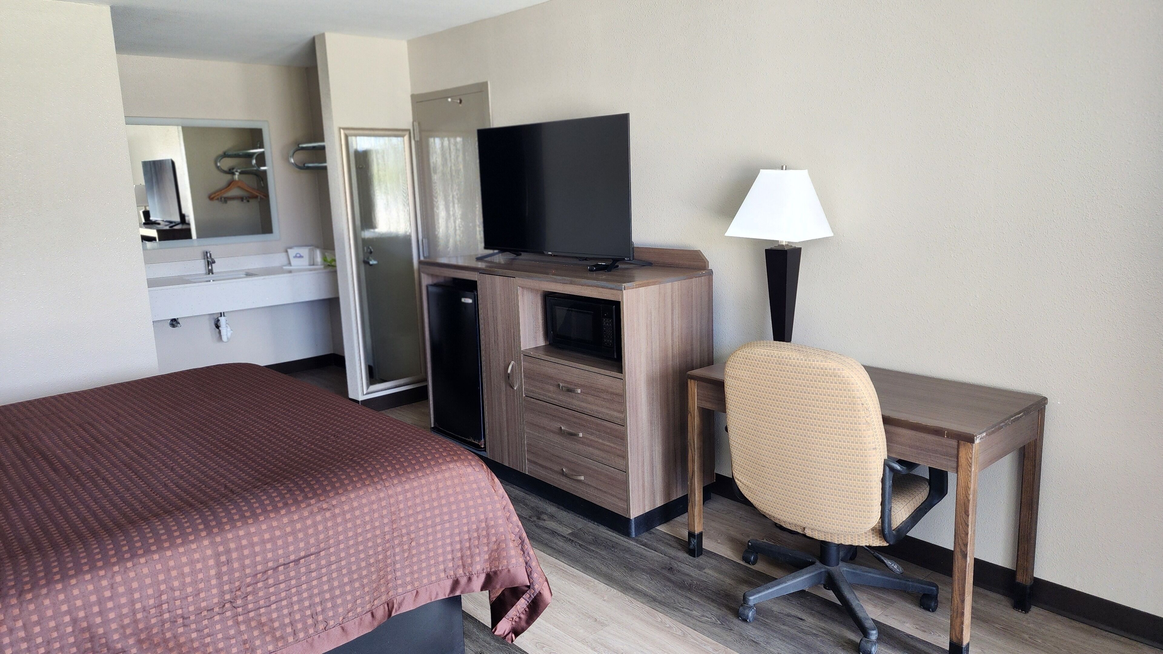 A hotel room with a bed, a desk with a chair, a television, a microwave, a refrigerator, and a mirror.