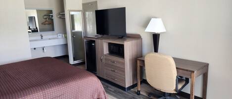 A hotel room with a bed, a desk with a chair, a television, a microwave, a refrigerator, and a mirror.