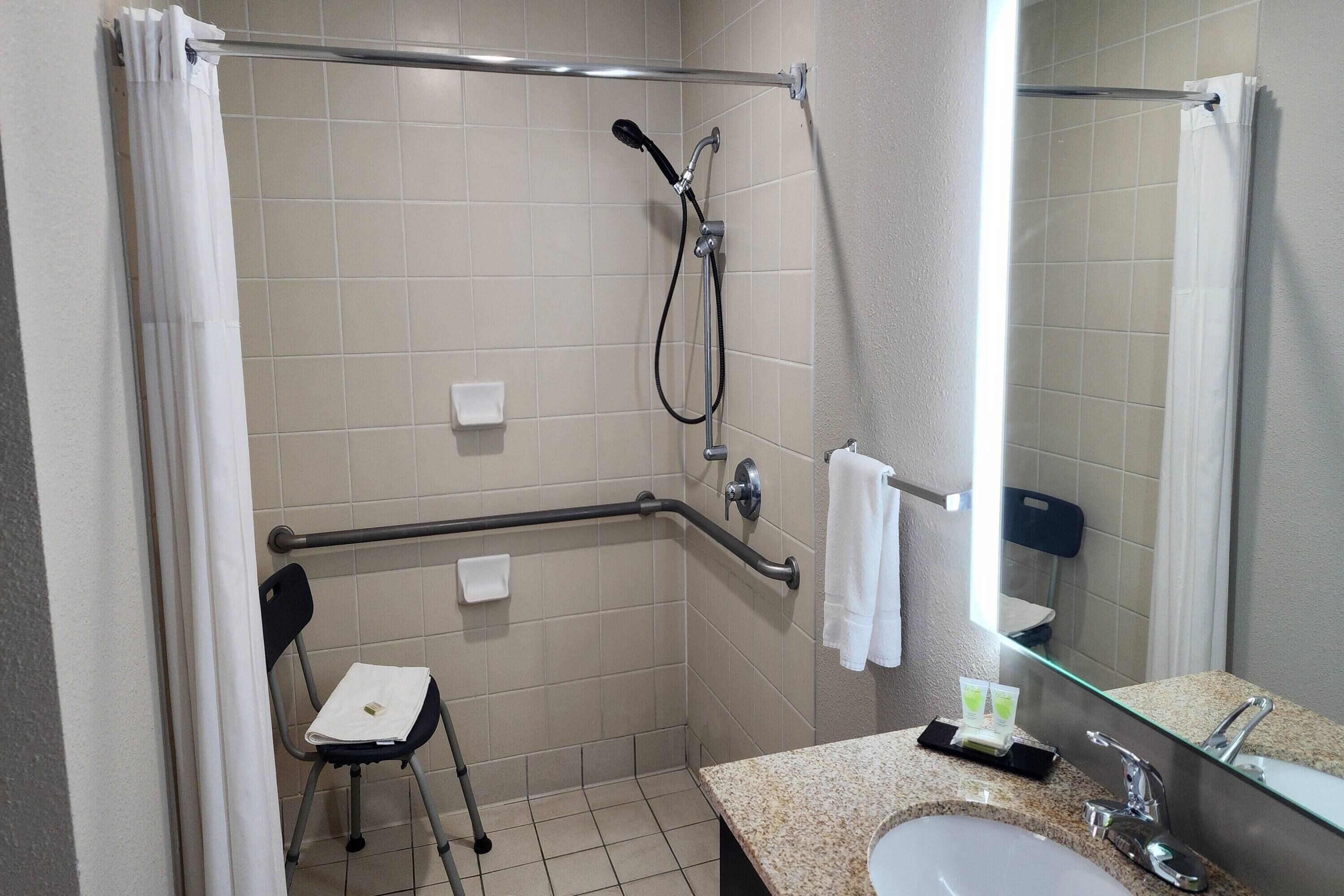 Baño con ducha, barra de apoyo, silla, lavabo y espejo.
