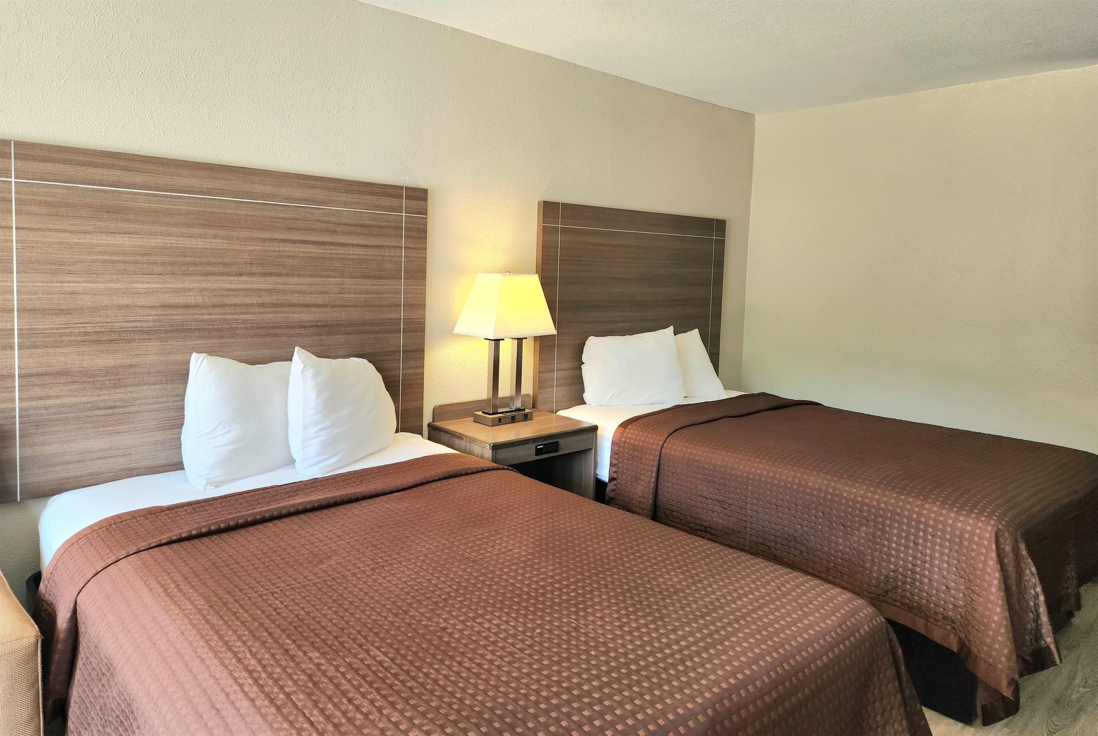 A hotel room with two beds, each with a brown bedspread, a nightstand with a lamp, and a headboard with a wooden panel design.