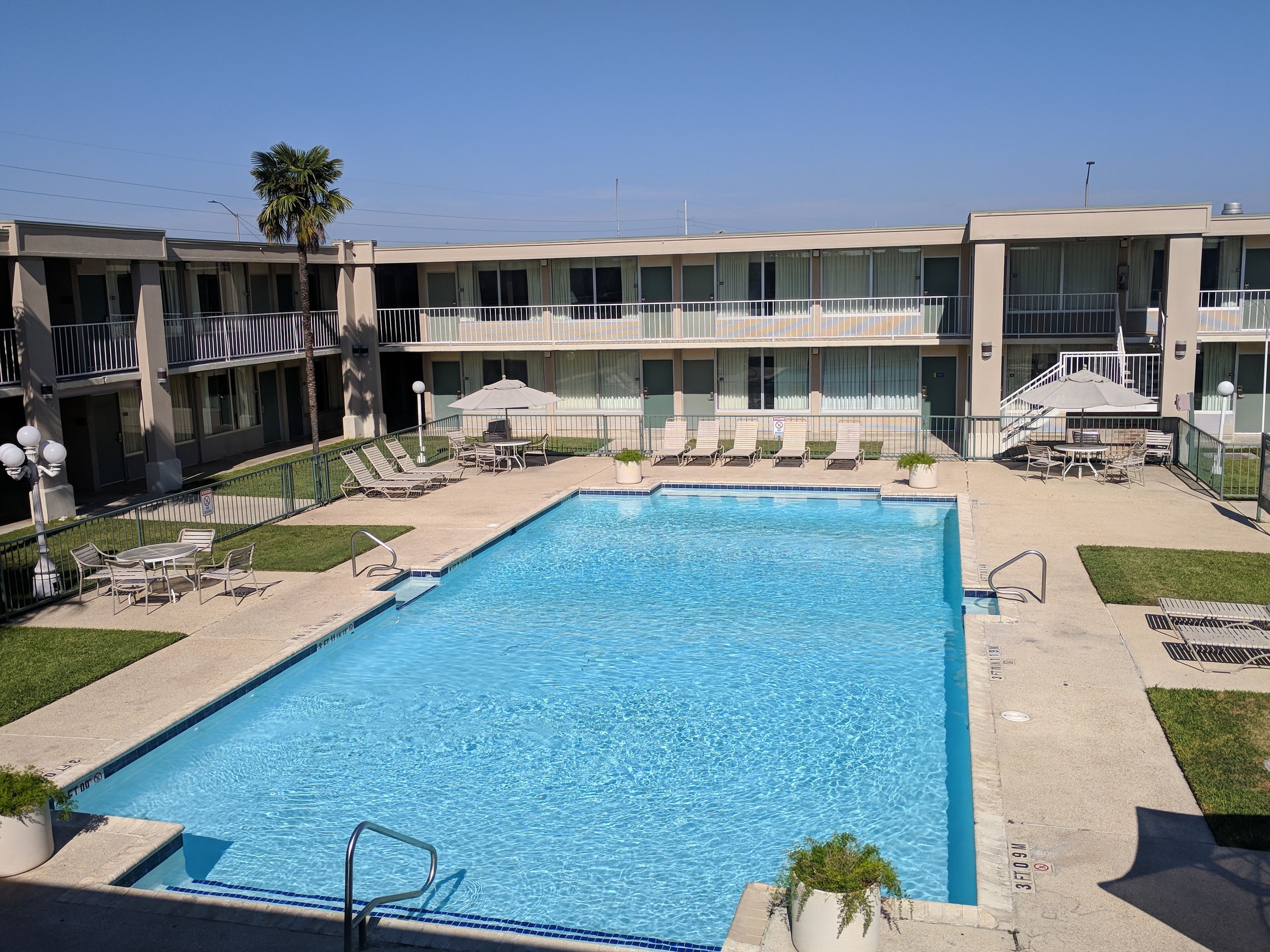 A rectangular pool with a tiled edge, surrounded by lounge chairs and umbrellas, with a building featuring balconies and a covered patio area in the background.