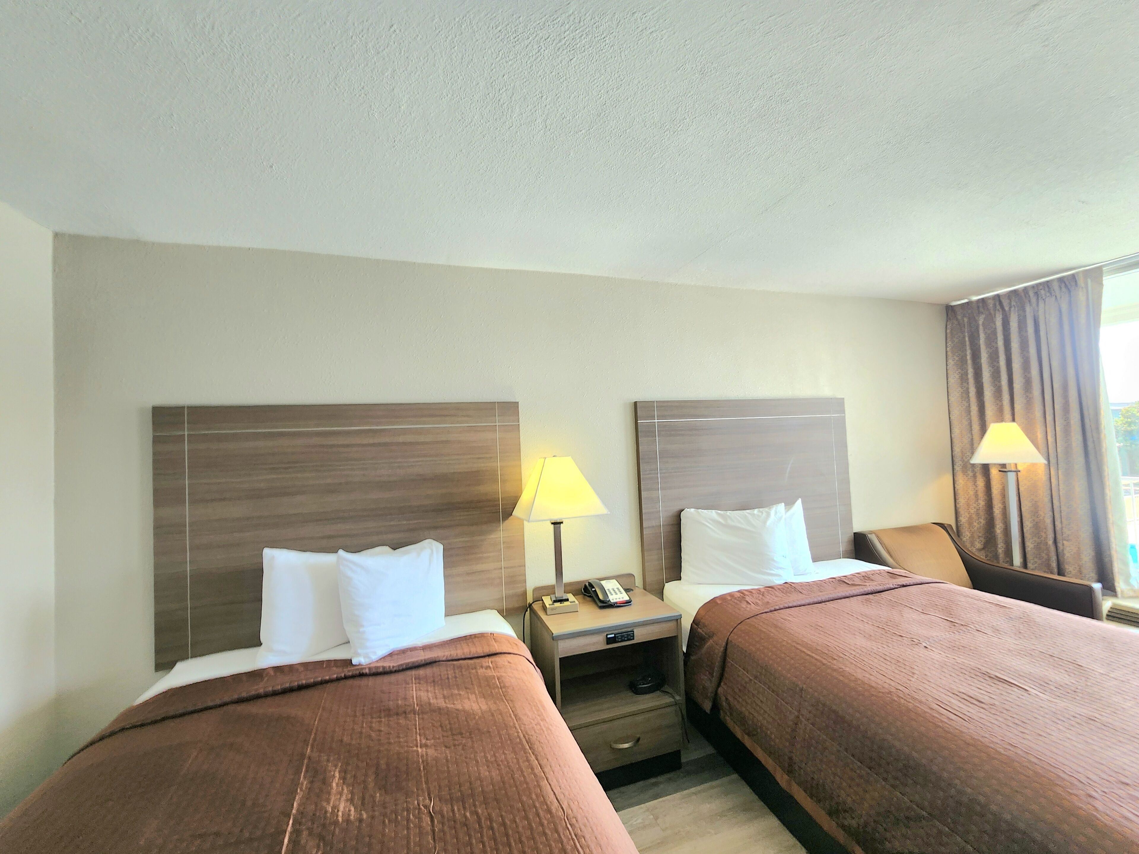 Habitación de hotel con dos camas, cada una con cabecero de madera, una mesita de noche con lámpara y un pequeño cajón.