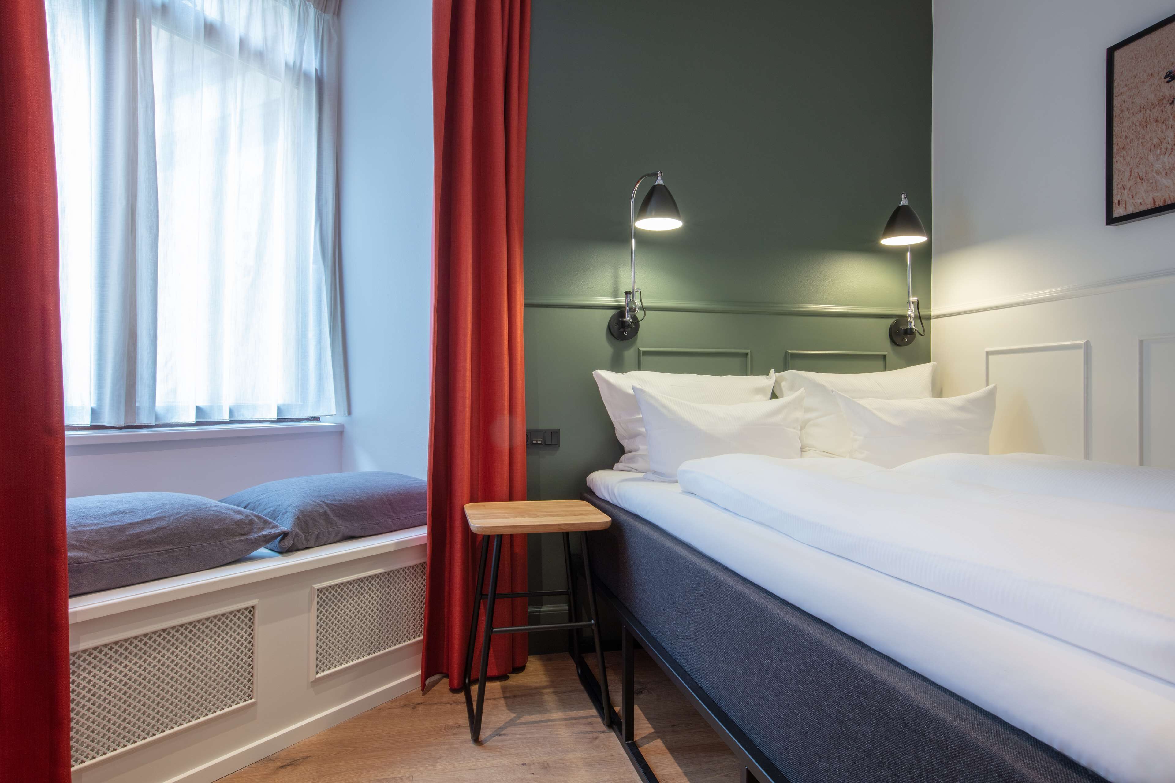 Un dormitorio con cama, ventana con cortinas, un taburete y dos lámparas fijadas a la pared.