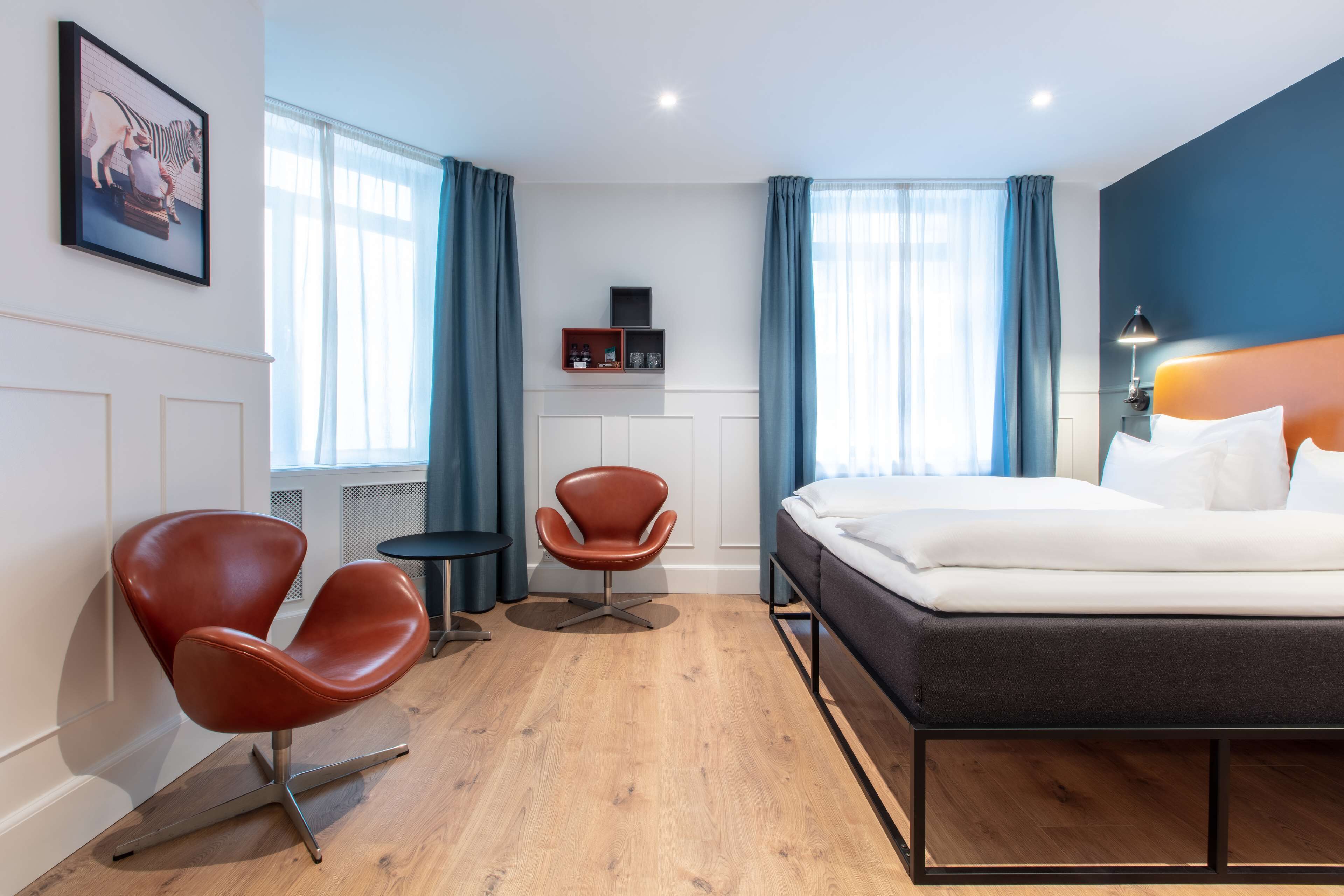 A modern hotel room with a bed, two chairs, a small table, and a shelf with items.